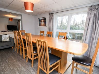 A dining room with a wooden table and chairs near large windows with curtains at Cadwaladr in Bontnewydd near Caernarfon