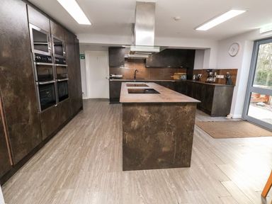 A kitchen with a large island, built-in ovens, cabinets, a coffee maker, and a door leading outside at Cadwaladr in Bontnewydd near Caernarfon