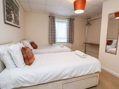 A bedroom with two single beds with white linens and plaid pillows a window with curtains a wall mirror and a hanging rail with hangers at Cadwaladr in Bontnewydd near Caernarfon