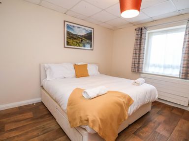 A bedroom with a double bed with white bedding and orange accents and a window with curtains at Cadwaladr in Bontnewydd near Caernarfon