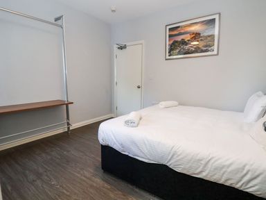 A bedroom with a bed with white linens two rolled towels on the bed a door a wooden shelf and a framed landscape picture on the wall at Cadwaladr in Bontnewydd near Caernarfon