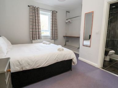 A bedroom with a bed, window with curtains, open wardrobe, wall mirror, and a view of an en suite bathroom at Cadwaladr in Bontnewydd near Caernarfon
