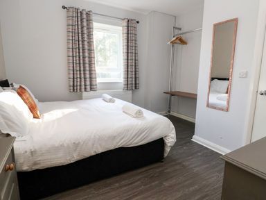 A bedroom with a bed with white linens two rolled towels a window with curtains a wall mirror and a clothing rack at Cadwaladr in Bontnewydd near Caernarfon