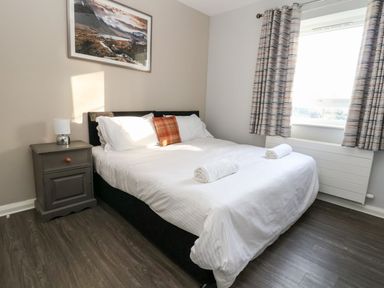 A bedroom with a double bed white bedding and two rolled towels on the bed beside a nightstand with a lamp and a window with patterned curtains at Cadwaladr in Bontnewydd near Caernarfon