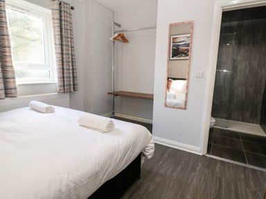 A bedroom with a bed, a window with curtains, a clothing rack with hangers, a wall mirror, and a bathroom entrance at Cadwaladr in Bontnewydd near Caernarfon