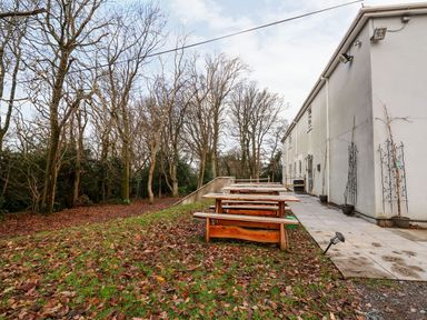 An outdoor area with wooden picnic tables next to a paved walkway and a white building surrounded by leafless trees at Cadwaladr in Bontnewydd near Caernarfon
