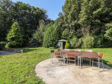An outdoor seating area with wooden chairs and tables on a paved surface surrounded by grass and trees at Cadwaladr in Bontnewydd near Caernarfon