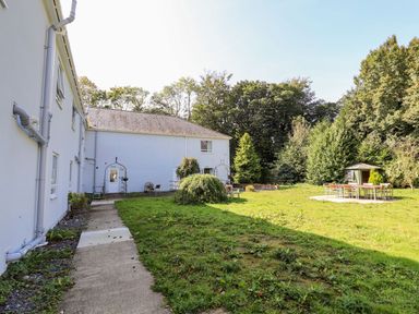 An outdoor area with a concrete pathway beside a white building a grassy lawn with scattered chairs a table set on a circular concrete slab and a small shed with trees in the background at Cadwaladr in Bontnewydd near Caernarfon