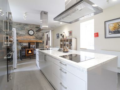 A kitchen with a large island countertop with stovetops and a kettle two range hoods cabinets a stone wall with a clock and a fireplace at The Lombard in Dolgellau