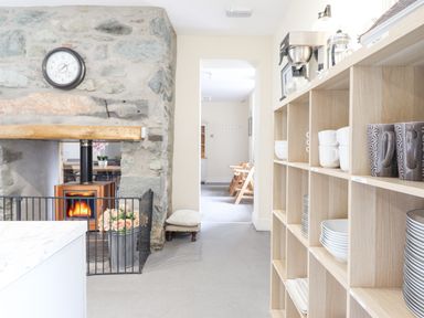 A kitchen area with a wooden shelving unit holding cups and plates a stone fireplace with a clock above and a fire burning in the stove at The Lombard in Dolgellau