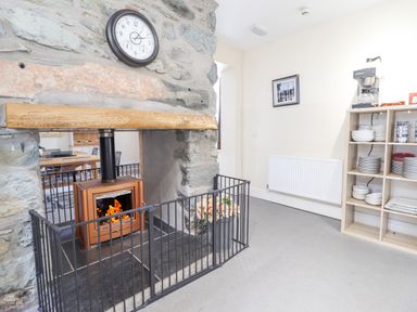 A stone fireplace with a wood burning stove and fireguard a wooden shelf with a clock above and shelving unit with dishes and coffee machine at The Lombard in Dolgellau