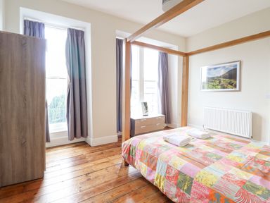 A bedroom with wooden floor a four-poster bed with a colorful quilt a chest of drawers two windows with curtains and a framed picture on the wall at The Lombard in Dolgellau