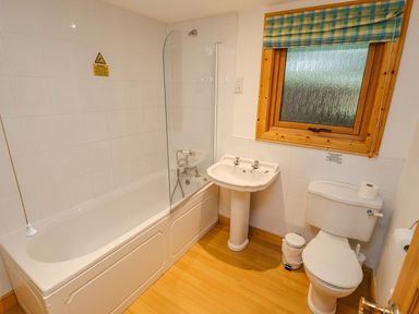 A bathroom with a bathtub and shower, a sink, a toilet, a window with a striped blind, and a small trash bin at Bamboo Retreat in Louth
