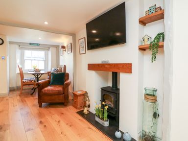 A living room with a leather armchair wooden floor small dining table under a window and a wood stove with a television above at Peppers Cottage in Appledore