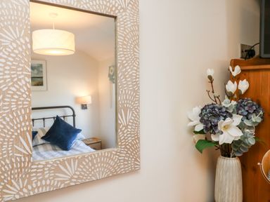 A bedroom reflection in a decorative wall mirror with a bed with blue pillows and a bedside table next to a vase with artificial flowers at Peppers Cottage in Appledore