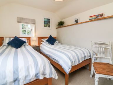 A bedroom with two single beds with striped bedding a wooden chair and a shelf with books and framed pictures at Pepper's Cottage in Appledore