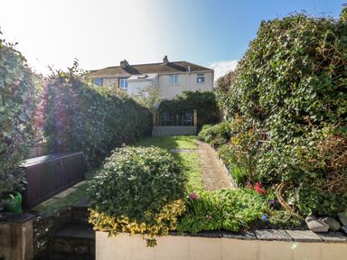 A backyard garden with a pathway leading to a deck with chairs surrounded by hedges and plants at Pepper's Cottage in Appledore