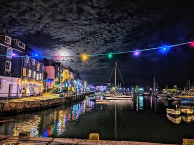 A night view of a harbor with boats docked and buildings with lights reflecting on the water at Mariners Rest in Gorran Haven