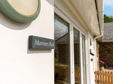 A sign reading Mariners Rest mounted on a white wall next to a round light above glass doors at Mariners Rest in Gorran Haven