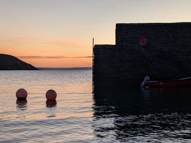 A waterfront with two buoys floating in water near a stone wall and a boat at Mariners Rest in Gorran Haven
