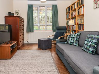 A living room with a grey couch with cushions a wooden bookshelf a blue armchair a wooden dresser and a TV at Greenhedges in Budleigh Salterton