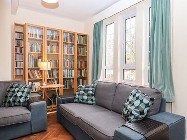 A sitting room with grey sofas patterned cushions a wooden bookcase with glass doors and windows with light blue curtains at Greenhedges in Budleigh Salterton