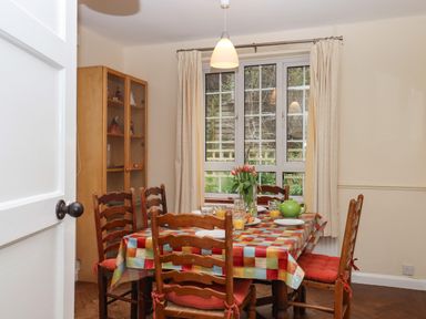 A dining room with a wooden table covered by a checkered tablecloth and six chairs near a window with cream curtains at Greenhedges in Budleigh Salterton