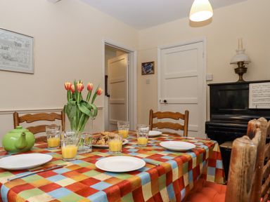 A dining room table set with glasses of orange juice plates cutlery a green teapot a vase of tulips near wooden chairs and a piano at Greenhedges in Budleigh Salterton