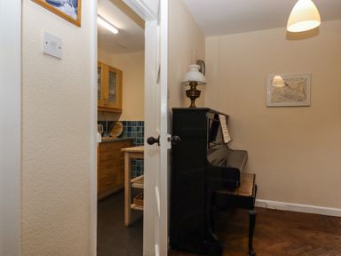 A black upright piano with sheet music and a lamp on top next to a door leading to a kitchen with wooden cabinets at Greenhedges Budleigh Salterton