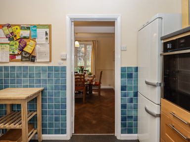 A kitchen with blue tiled walls a wooden table fridge and oven looking through a doorway to a dining room with a table and chairs at Greenhedges in Budleigh Salterton