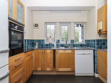 A kitchen with wooden cabinets blue tiled backsplash white dishwasher and a double sink below three windows at Greenhedges in Budleigh Salterton