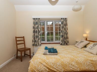 A bedroom with a double bed covered with a yellow floral duvet a wooden chair a window with patterned curtains and a bedside table with a lamp at Greenhedges Budleigh Salterton