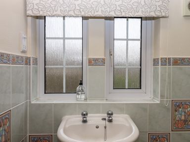 A bathroom sink with two taps and a soap dispenser under a window with floral patterned tiles at Greenhedges in Budleigh Salterton