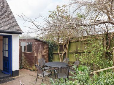 An outdoor patio area with a round metal table and five chairs near a wooden shed and a green wooden fence with some plants at Greenhedges in Budleigh Salterton