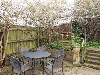 An outdoor patio area with a round metal table and four metal chairs surrounded by trees a wooden fence and a gate at Greenhedges in Budleigh Salterton