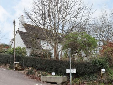A white house with a steep roof behind leafless trees and bushes at a roadside junction with road signs and a convex mirror in Budleigh Salterton