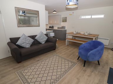 A living room with a dark sofa, blue armchair, patterned rug, dining table with bench, and kitchen area at 16 Coedrath Park in Saundersfoot