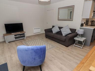 A living room with a blue chair a gray sofa with cushions a TV on a stand and a side table with a lamp at 16 Coedrath Park in Saundersfoot