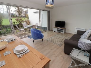 A living room with a brown sofa a blue chair a TV on a stand and a dining table set with dishes at 16 Coedrath Park in Saundersfoot