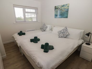 A bedroom with two beds with white bedding green towels and blue patterned pillows a nightstand with a lamp and a window with blinds at 16 Coedrath Park in Saundersfoot