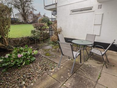 An outdoor patio with a round glass table and four chairs next to a garden with plants and grass at 16 Coedrath Park in Saundersfoot