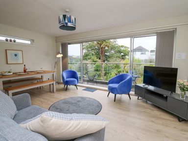 A living room with blue armchairs a sofa a TV on a cabinet a dining table and sliding glass doors at 36 Coedrath Park in Saundersfoot