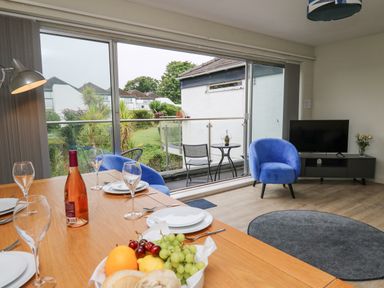 A dining area with table set with plates and wine glasses next to a seating area with blue chairs and a TV at 36 Coedrath Park in Saundersfoot