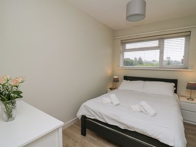 A bedroom with a bed with white bedding and towels on it a window with blinds two bedside tables with lamps and a white dresser with a vase of flowers at 36 Coedrath Park Saundersfoot