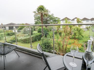 A balcony with two chairs a round table with two wine glasses and a bottle overlooking a garden with palm trees at 36 Coedrath Park in Saundersfoot