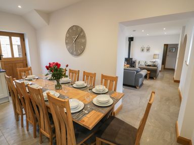 A dining room with a wooden table set with plates and glasses and a clock on the wall next to a living room with sofas and a wood stove at The Stables in Droitwich Spa
