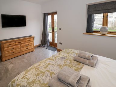 A bedroom with a wooden dresser a wall-mounted TV folded towels on a bed and glass doors leading outside at The Stables in Droitwich Spa