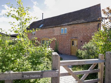 A brick house with wooden doors and windows behind a wooden gate with green plants at The Stables in Droitwich Spa