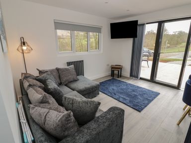 A living room with a gray sectional sofa a blue rug a wall-mounted tv and glass doors leading outside at Goldthorne Lodge in Clee Hill