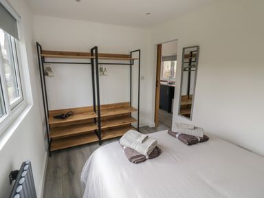 A bedroom with a bed and folded towels on it open wooden shelves with hangers a window and an open door to a kitchen at Goldthorne Lodge in Clee Hill